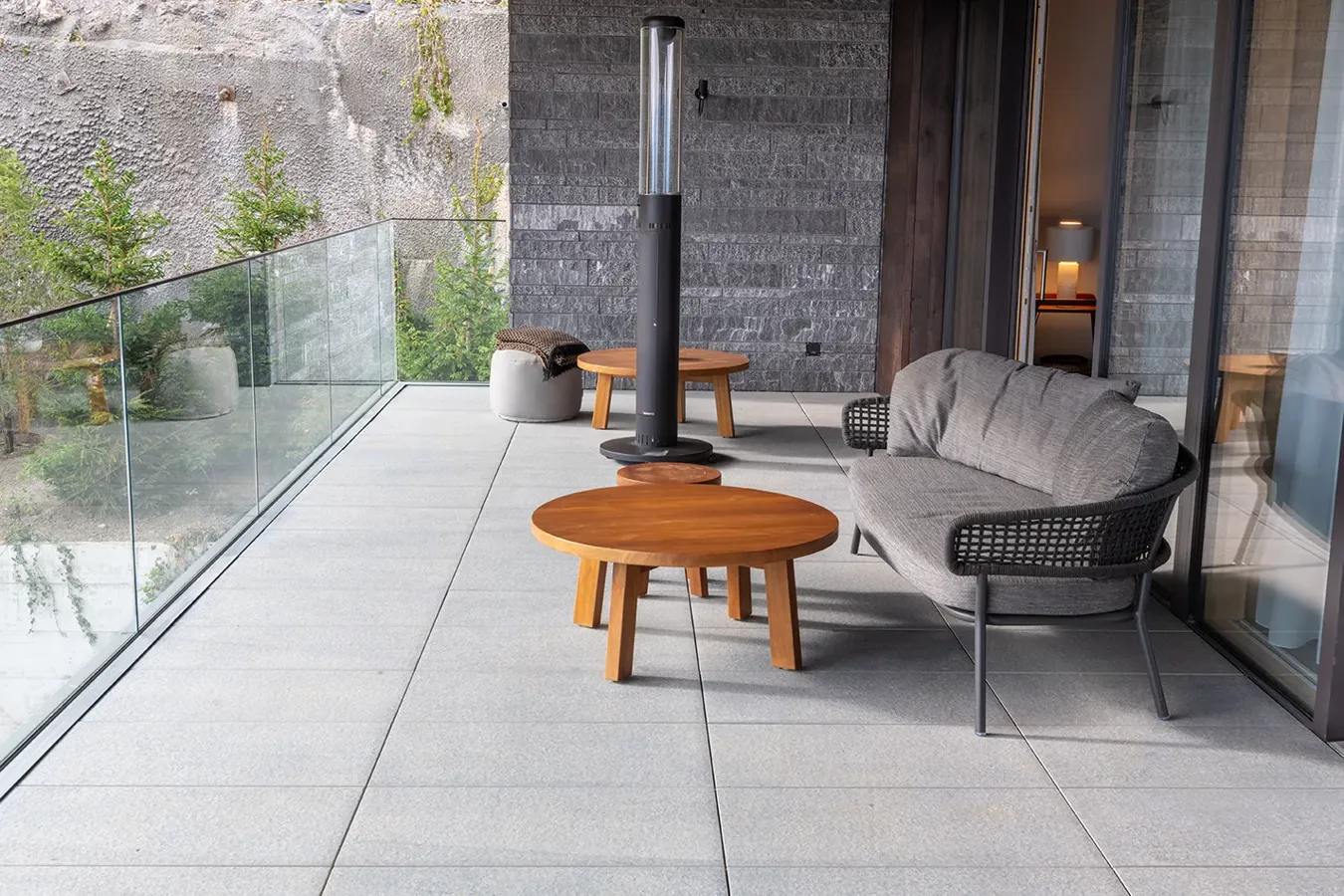 Stilvolle Terrasse mit grauen Feinsteinzeugfliesen und Außenmöbeln aus Stoff und Holz, umgeben von Steinwänden und Glas.