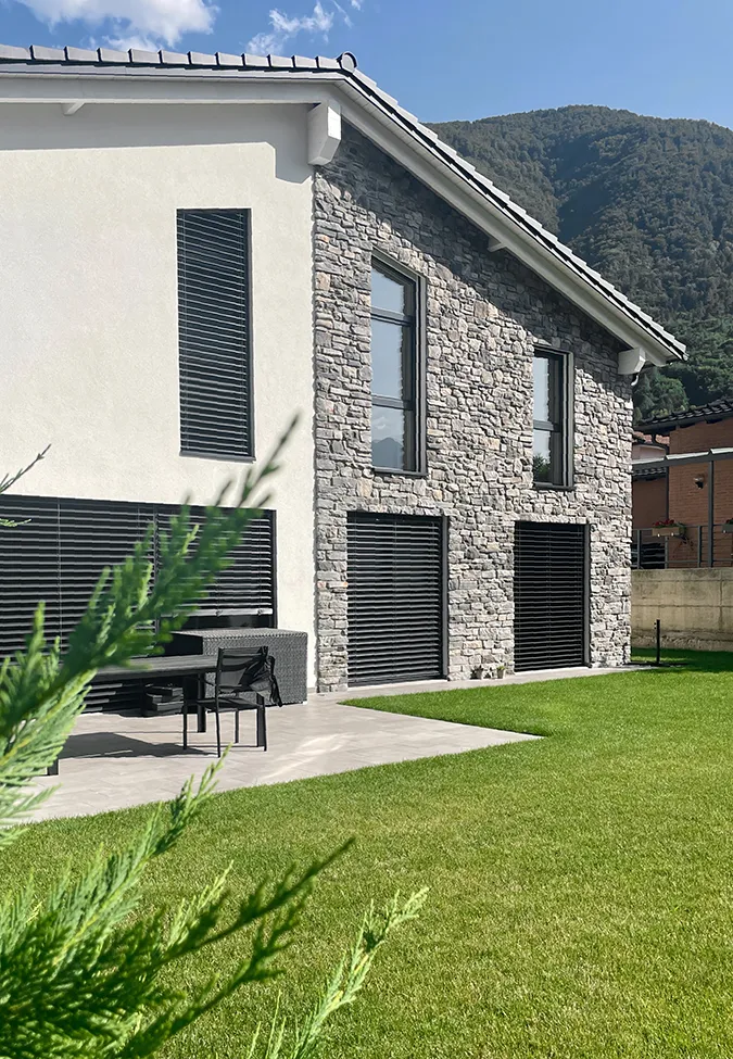 Esterno di casa in montagna con piastrelle effetto pietra grigio della collezione Noord, prato verde e mobili da giardino.