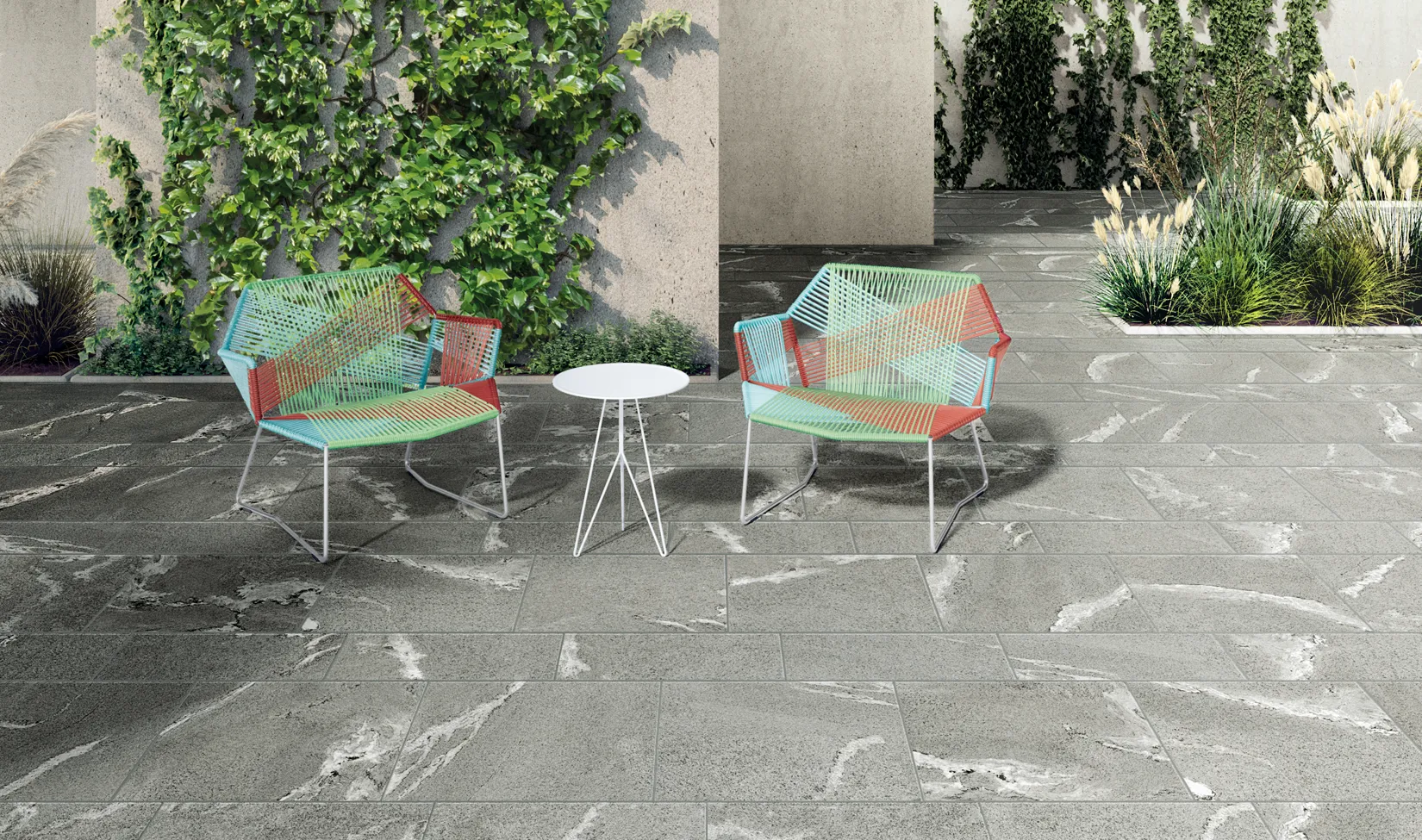 Patio esterno con piastrelle effetto pietra della Percorsi Frame colore Gneiss Grey, abbinato a sedie a rete colorate e tavolino rotondo, circondato da giardini pietrificati e vegetazione.