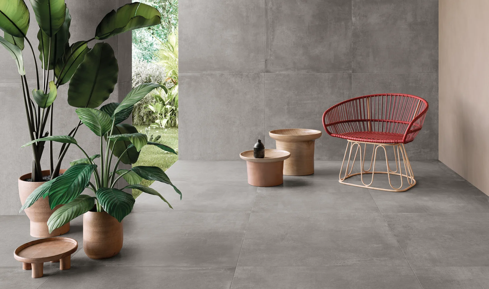 Interni moderni con pavimento in gres porcellanato grigio chiaro della collezione Noord tonalità Grey, arredati con una sedia in filo rosso e tavolini in terracotta, e piante verdi con sfondo di giardino esterno.