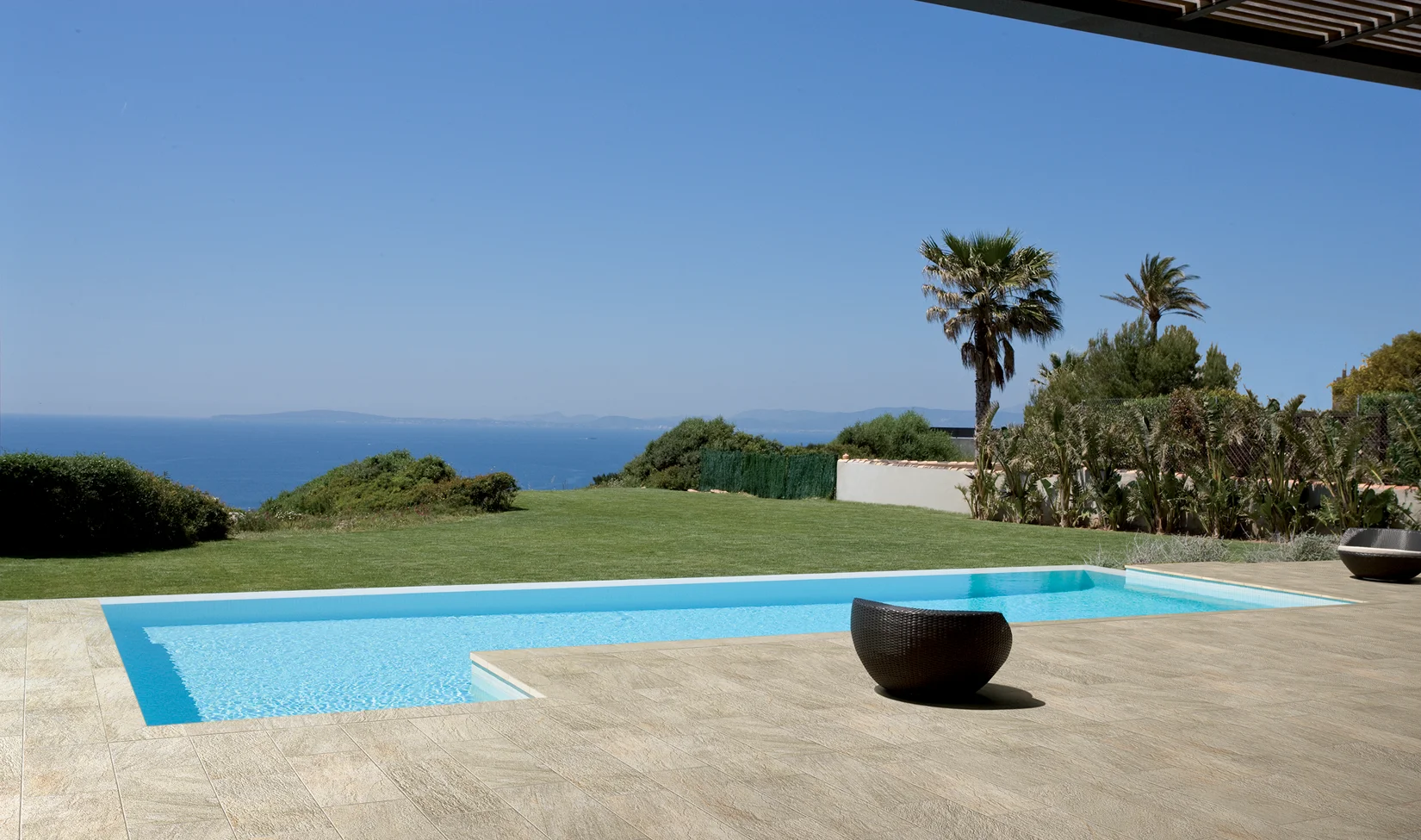 Piscina esterna con bordo in piastrelle effetto pietra, circondata da un giardino lussureggiante con palme e vista panoramica sul mare.