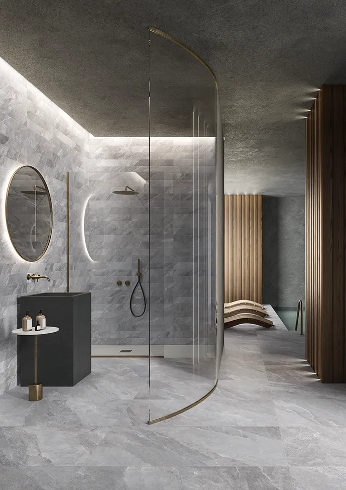 Интерьер шикарной ванной комнаты с серым керамогранитом коллекции Ubik, золотыми акцентами и криволинейной стеклянной душевой.