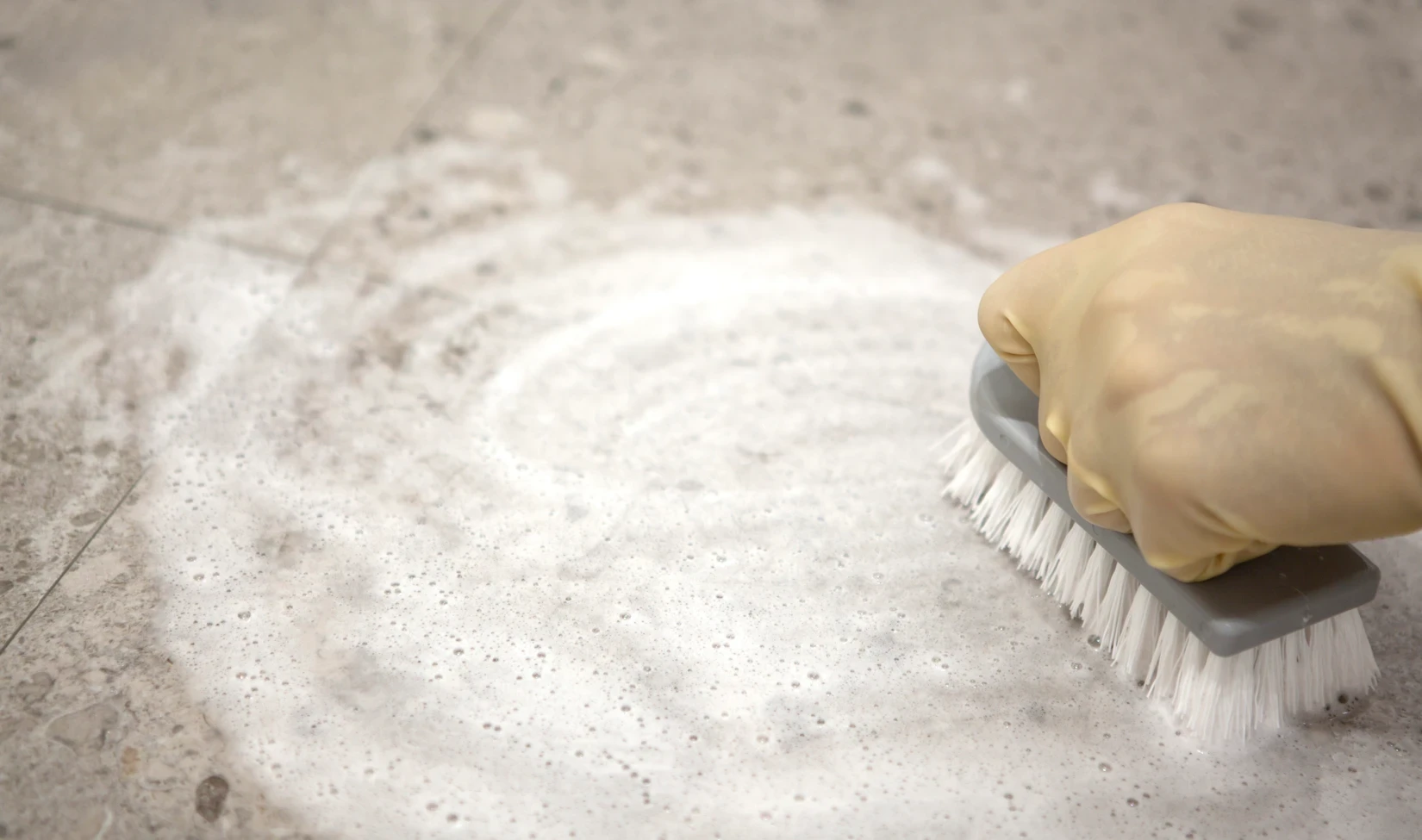 Hand in gelbem Schutzhandschuh schrubbt Feinsteinzeugboden mit schaumigem Reinigungsmittel und Bürste, Konzept der Fliesenbodenpflege.