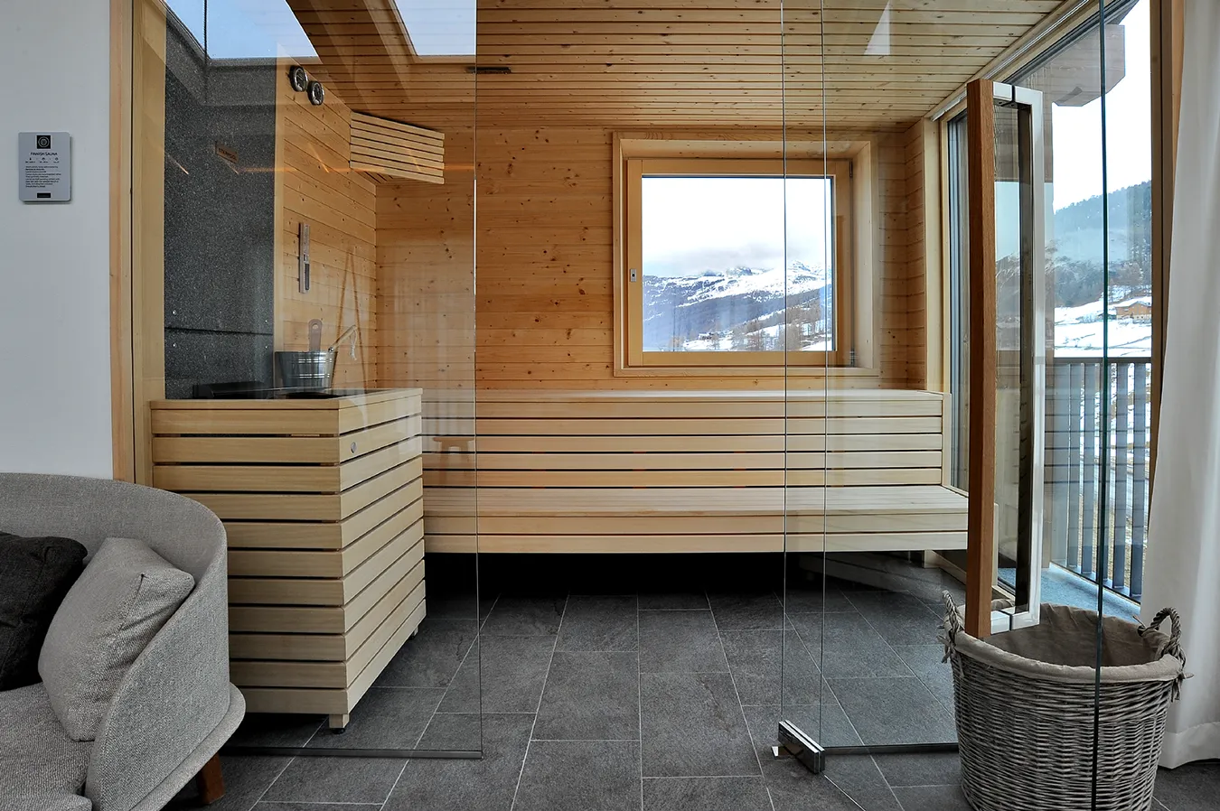 Elegant wooden sauna room with stone-effect porcelain stoneware flooring from the Percorsi Extra collection in Pietra di Combe color, offering mountain views through the window.