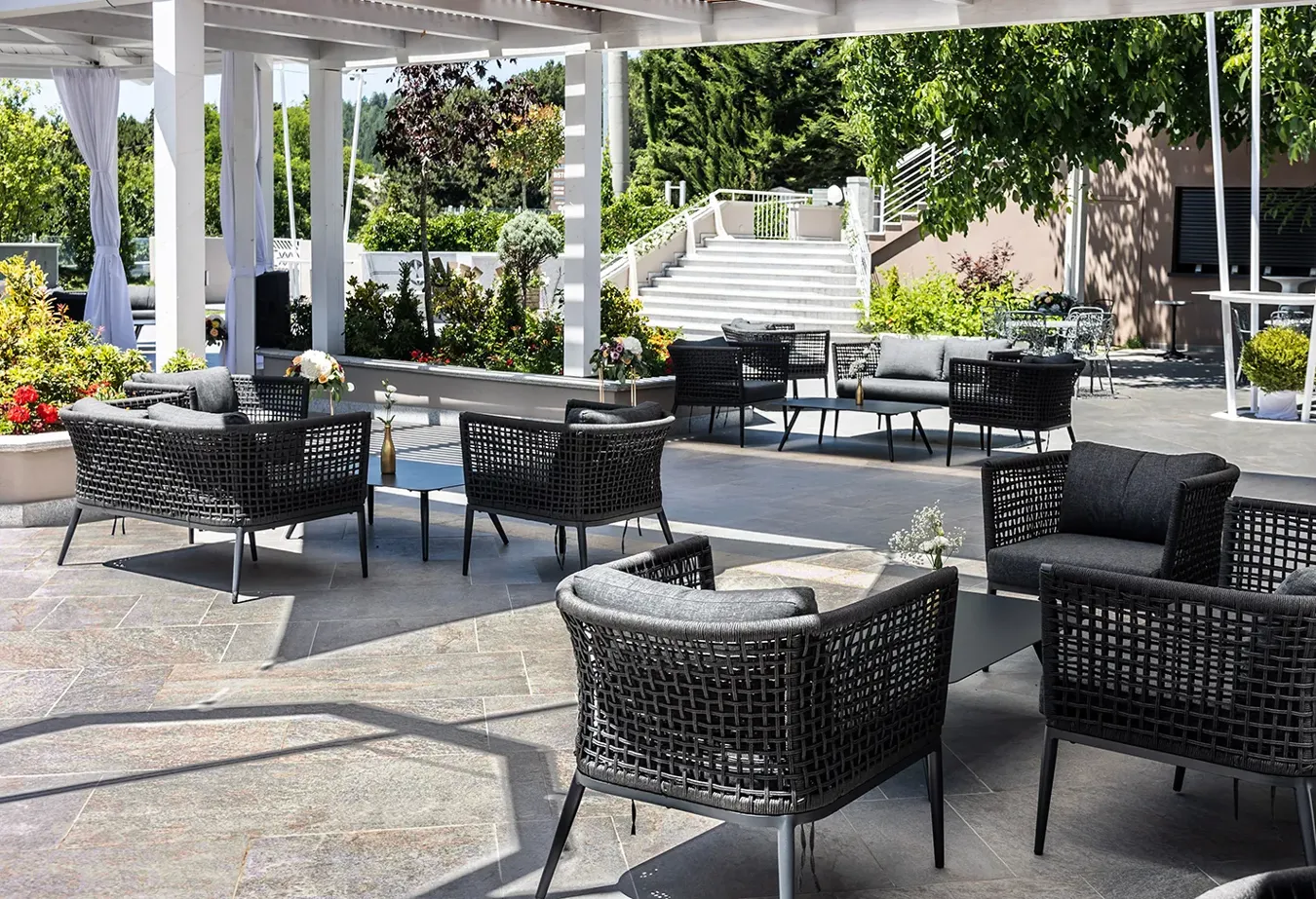 Sophisticated outdoor setting with Percorsi Extra stone effect tiles, black rattan furniture, and lush greenery.