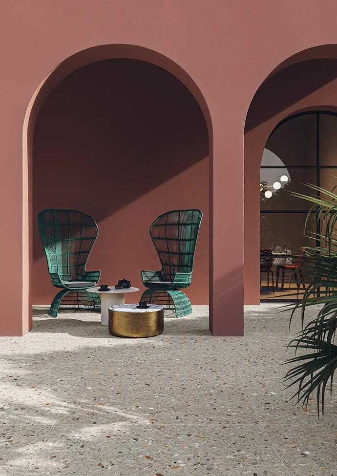 Piastrelle da esterno effetto pietra con sedie in filo verde e tavolino dorato sotto archi terracotta.