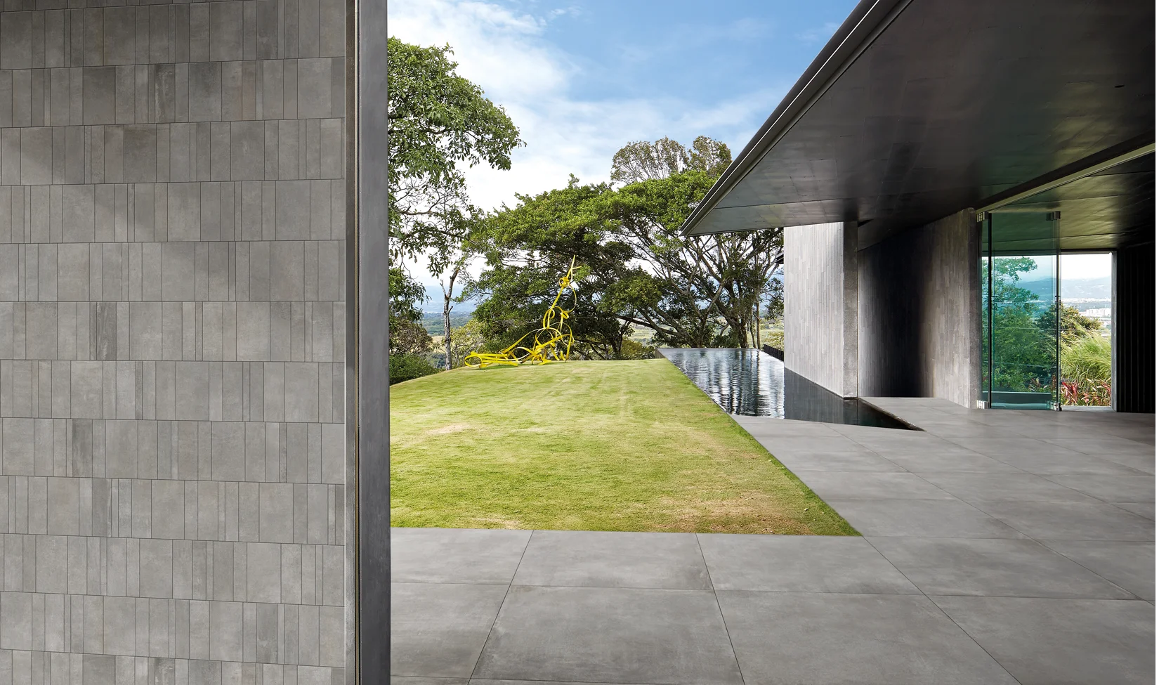 Un pavimento esterno in gres porcellanato effetto cemento Noord Grey, spessore 20 mm, con vista su un giardino e piscina a sfioro.