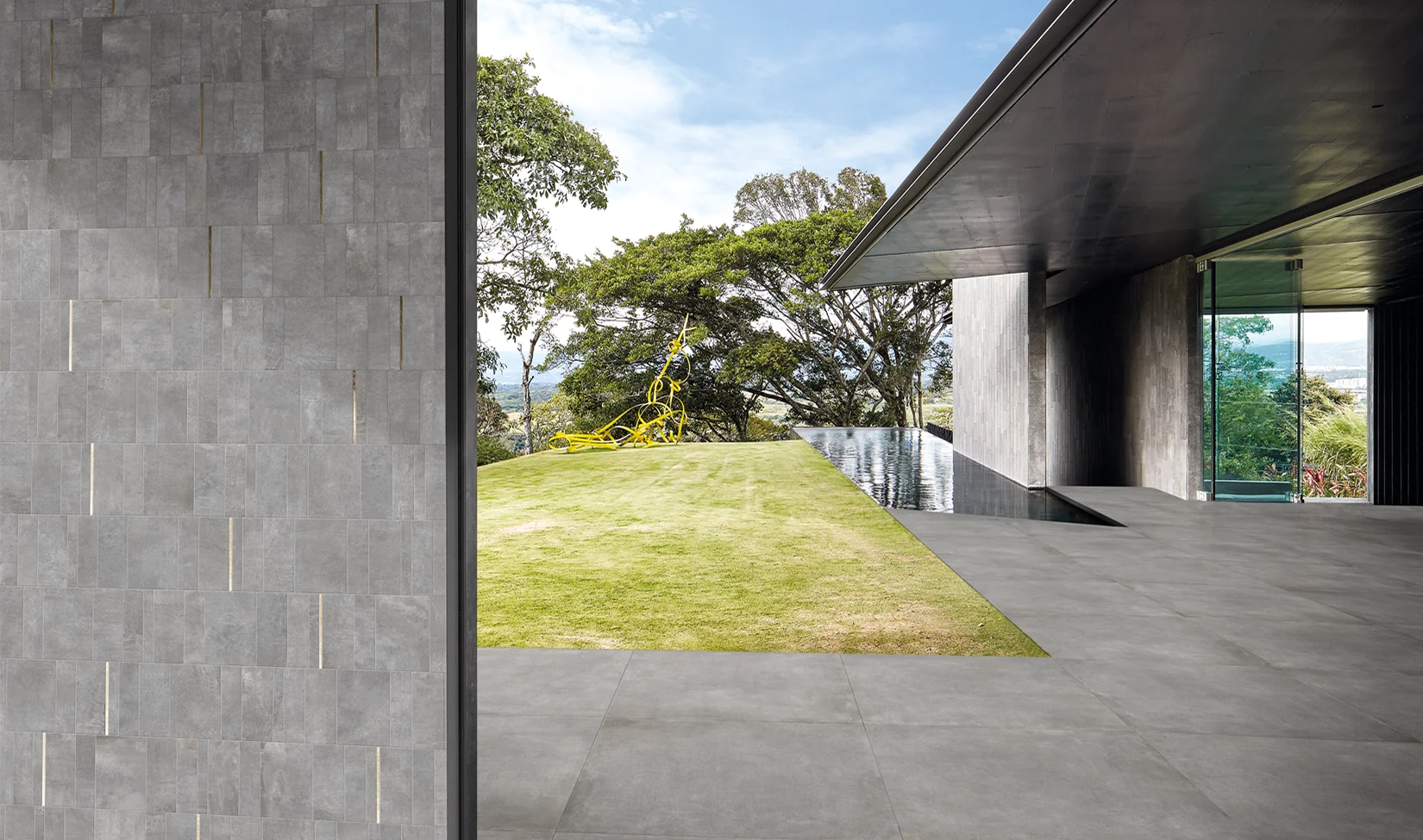 Ambiente moderno con pavimenti in gres porcellanato effetto cemento della collezione Noord colore grigio (Grey), vetrate ampie e continuità visiva verso un giardino esterno curato.